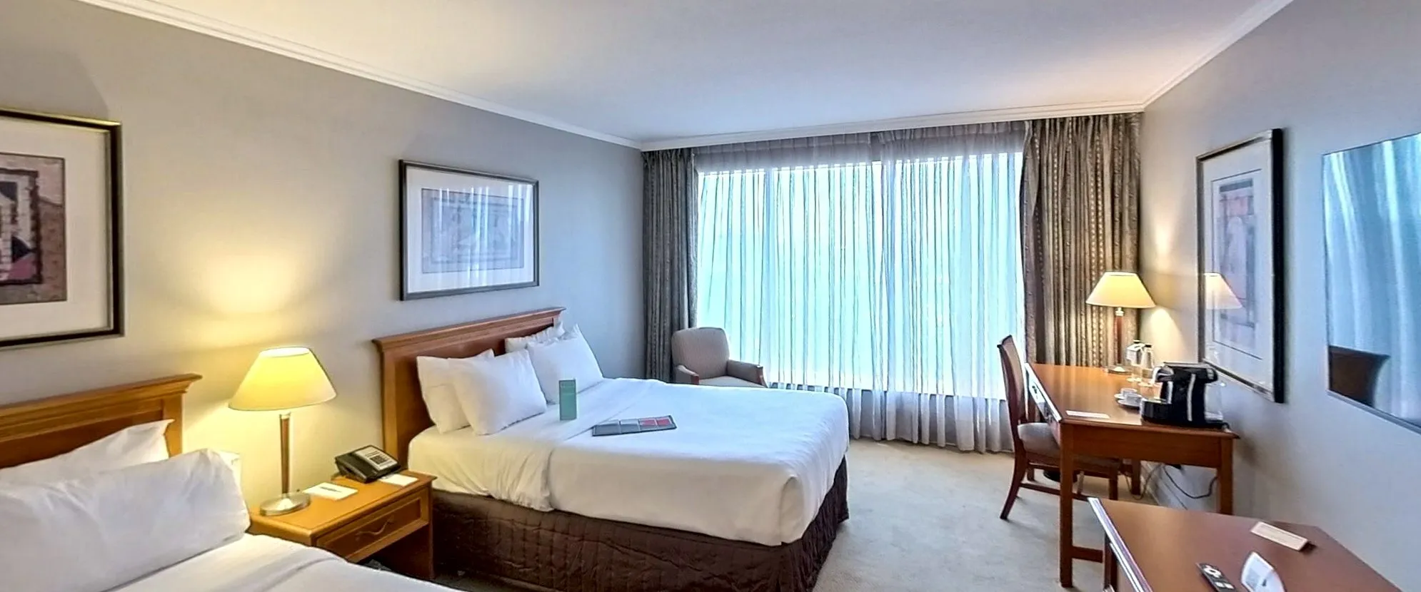A twin hotel room: two beds with white linen, lamps on the bedside table and the city through the window